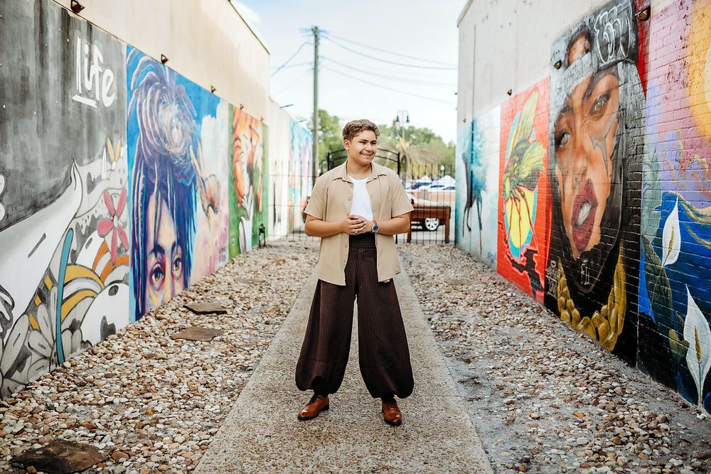 Young man stands in colorful mural alley wearing wide-leg pants and beige shirt, holding phone.