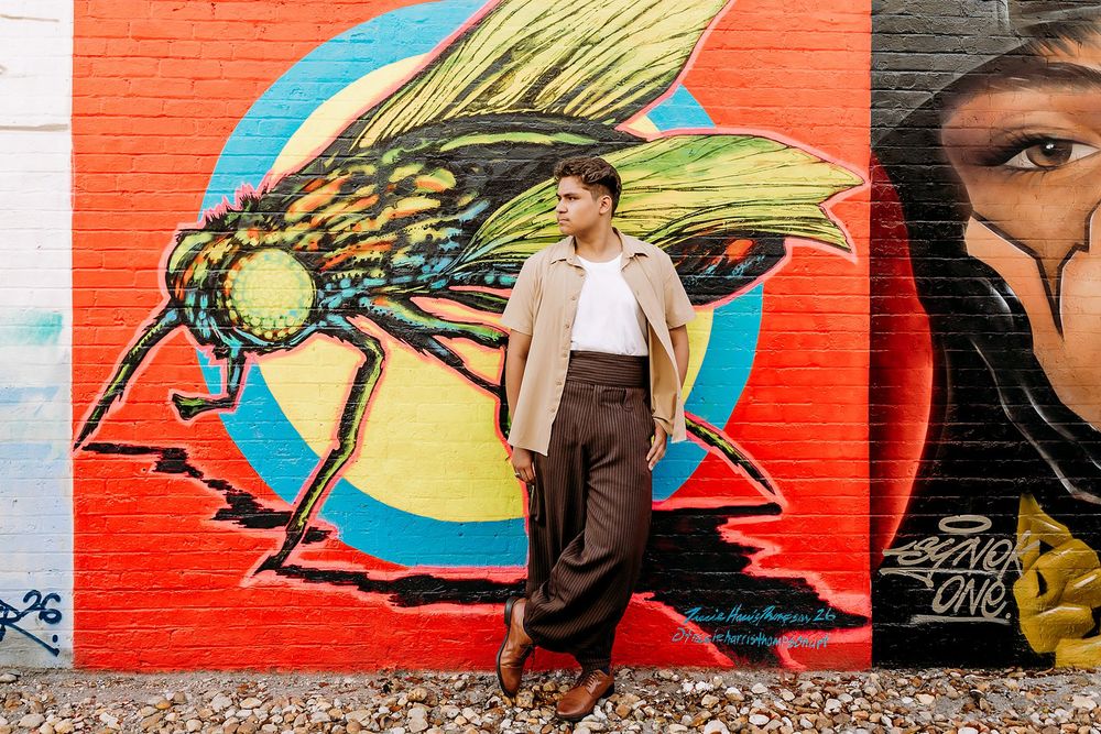 Person posing in front of vibrant street art mural featuring a large fly and portrait on a red brick wall.