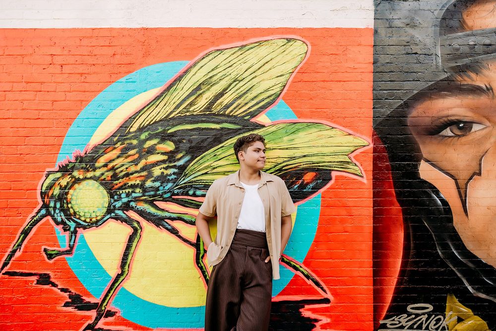 Young man posing in front of colorful street art mural featuring a large fly and portrait.