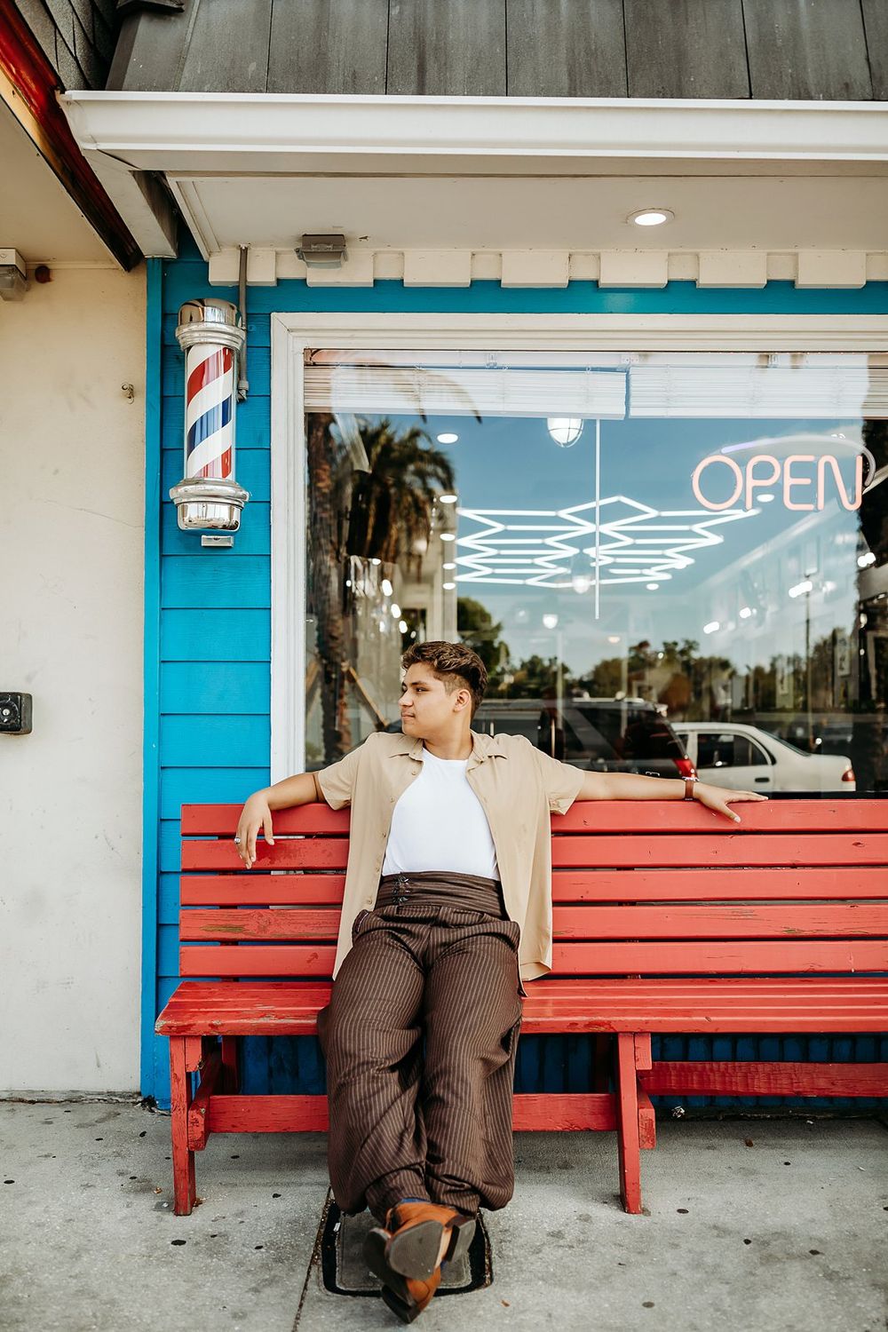 Young man relaxing on a red bench outside a barbershop with a barber pole and neon open sign.