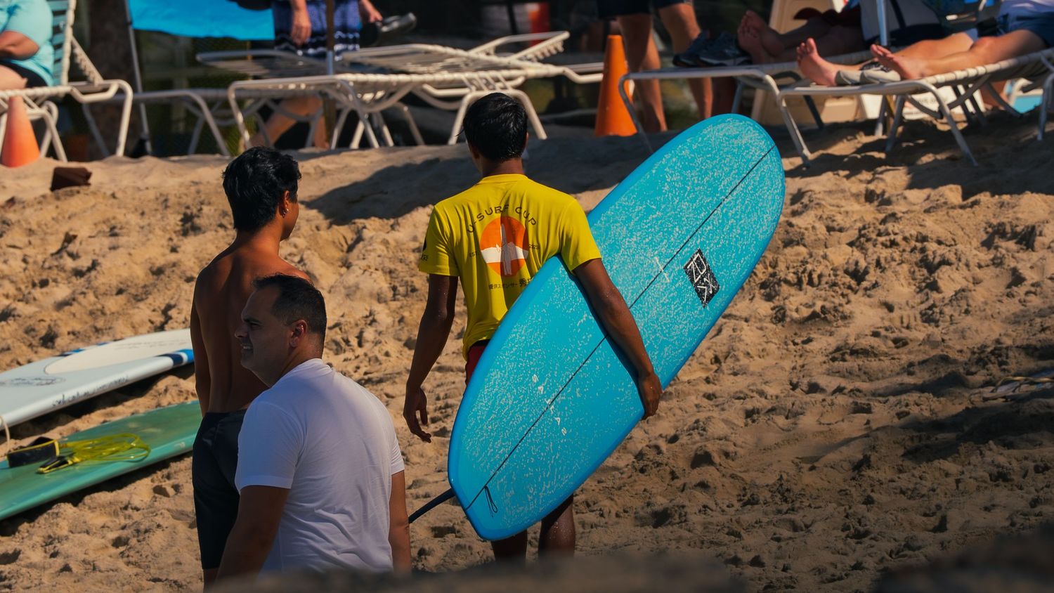 A surfer in a U Surf Cup shirt carries a blue surfboard across the sand at Waikiki as two others talk nearby at the beach event.