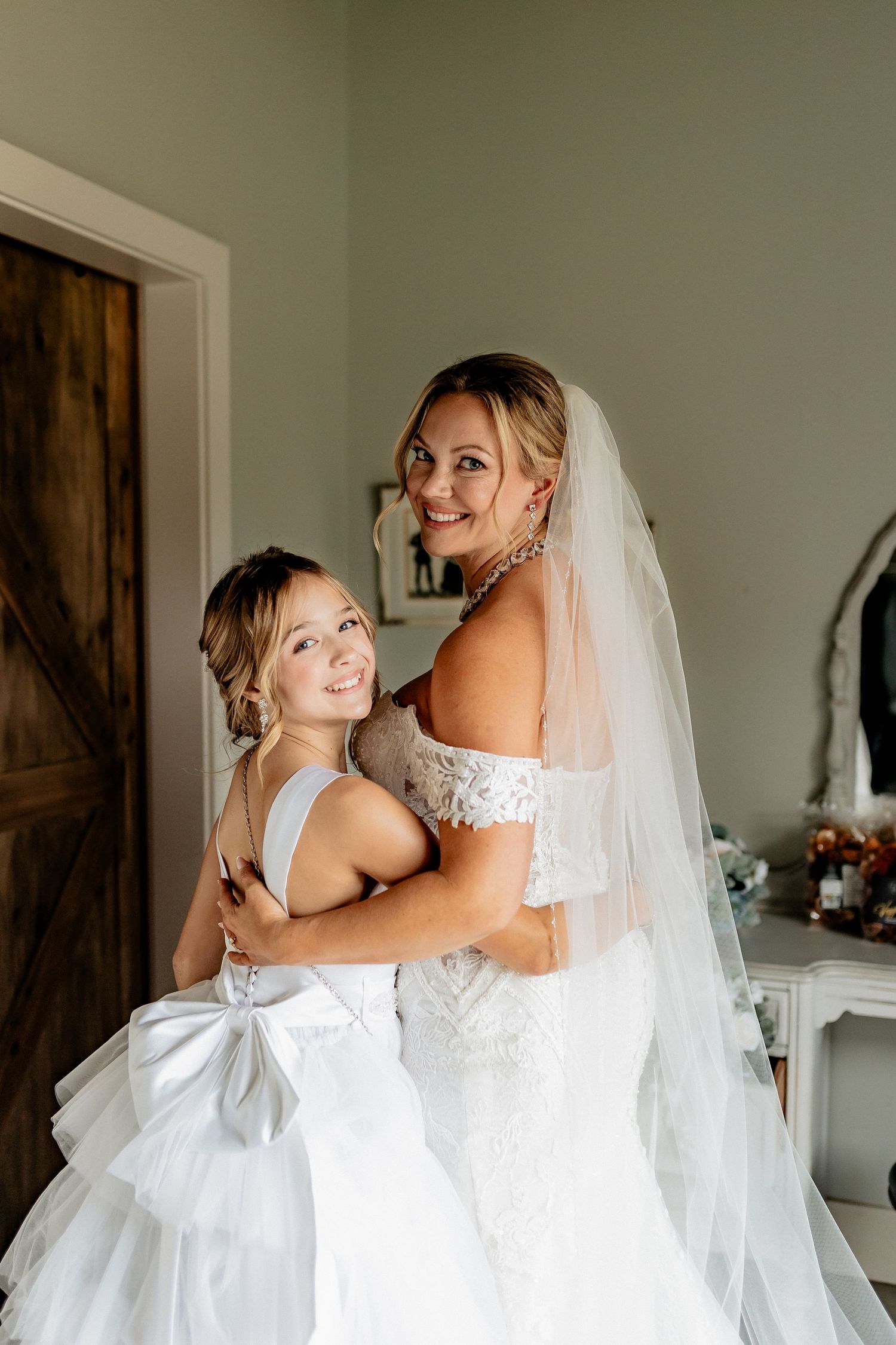Two people in white wedding dresses embrace while smiling at the camera in a bright room with a rustic door. Hawthorne Estates