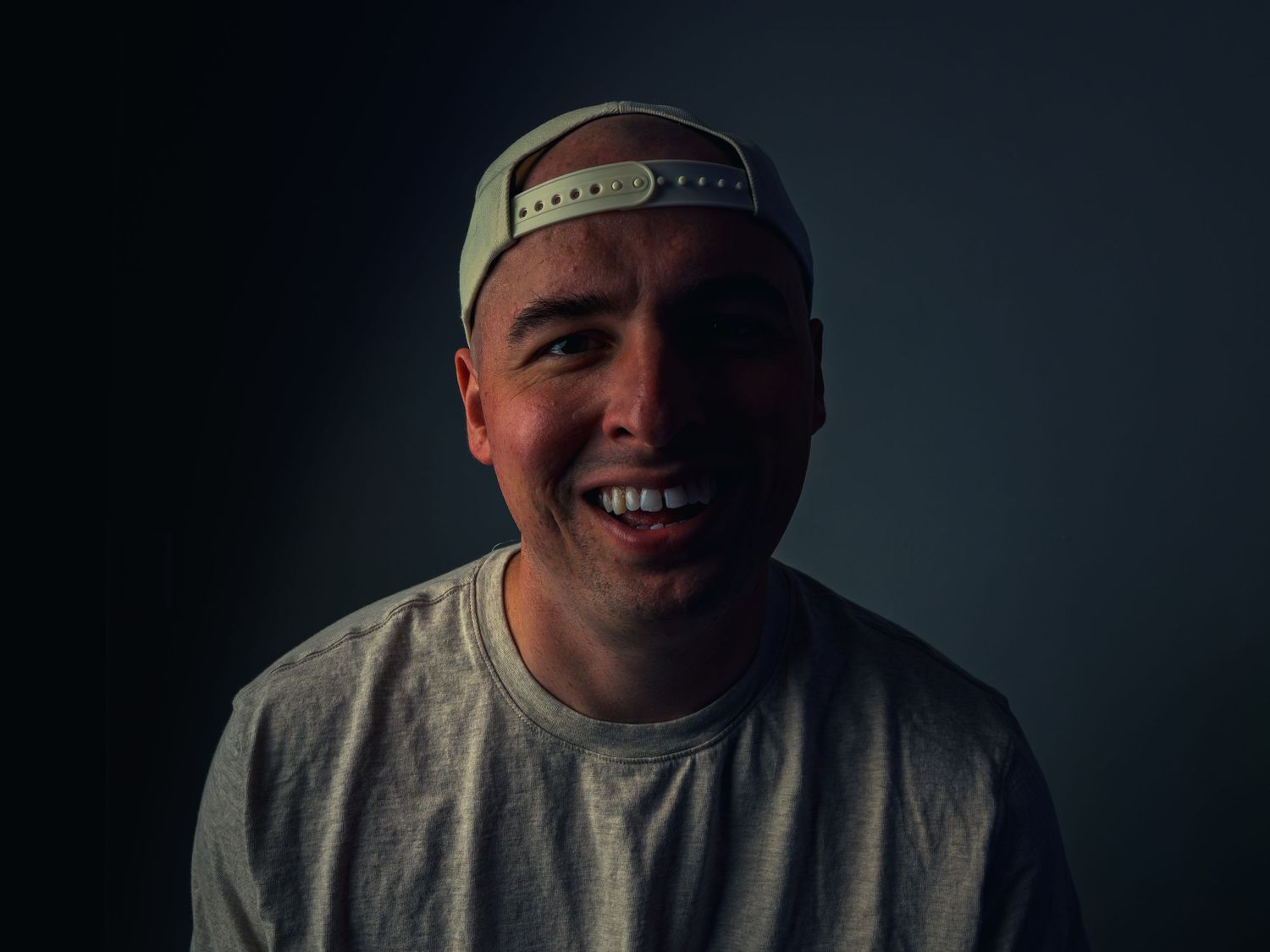 A man in a backward cap and gray tee smiles warmly straight at the camera, face lit by soft warm light against a dark teal background