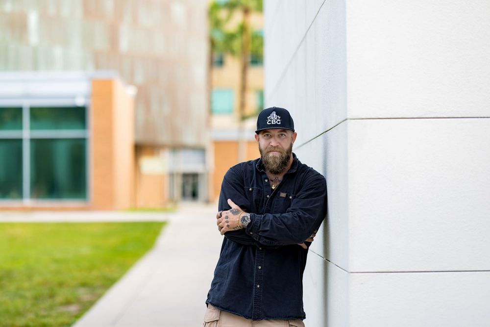 Man in dark jacket and cap leans against modern building wall outdoors on sunny day.