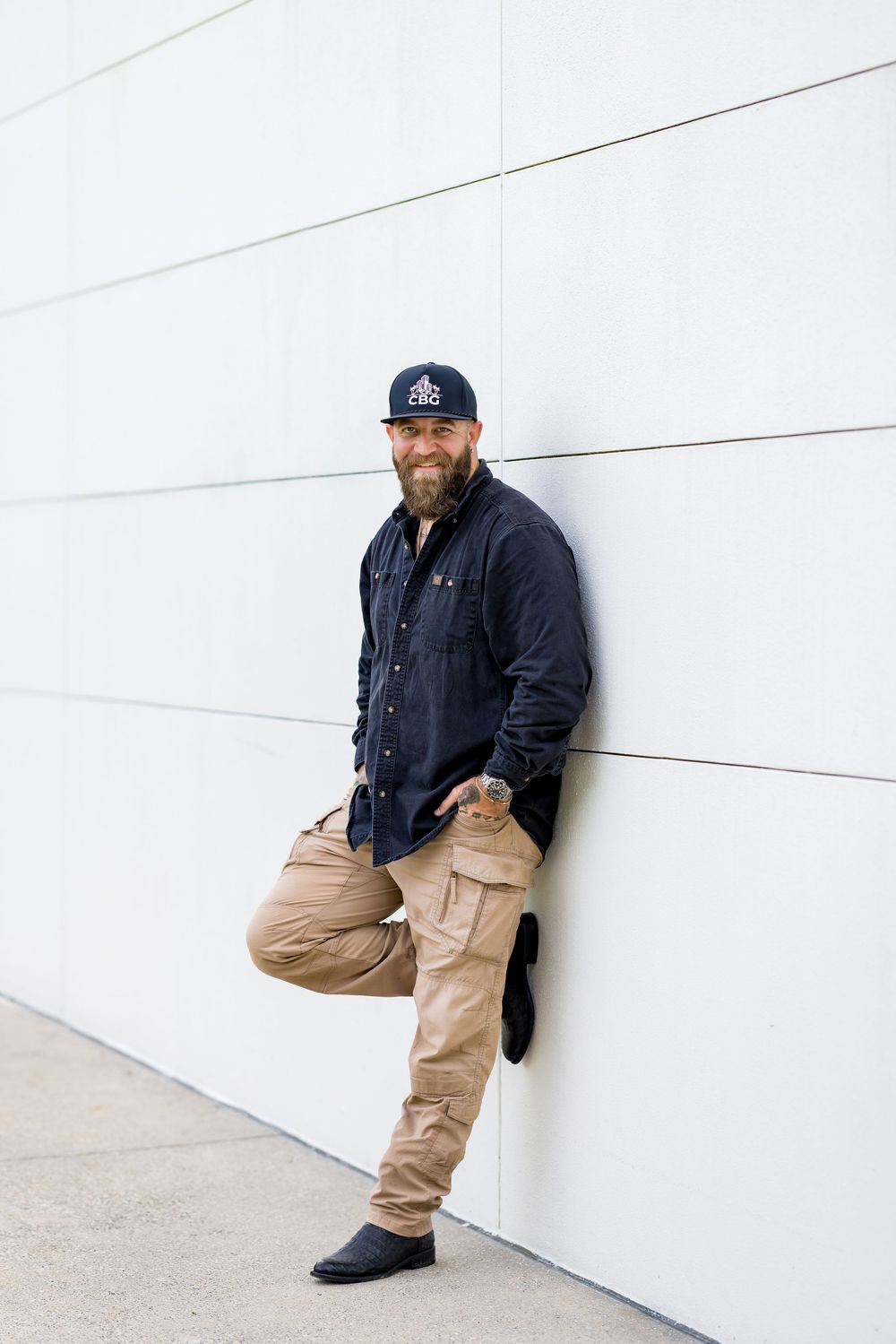 Bearded man in navy shirt, khaki cargo pants, and baseball cap leaning against a white wall smiling.