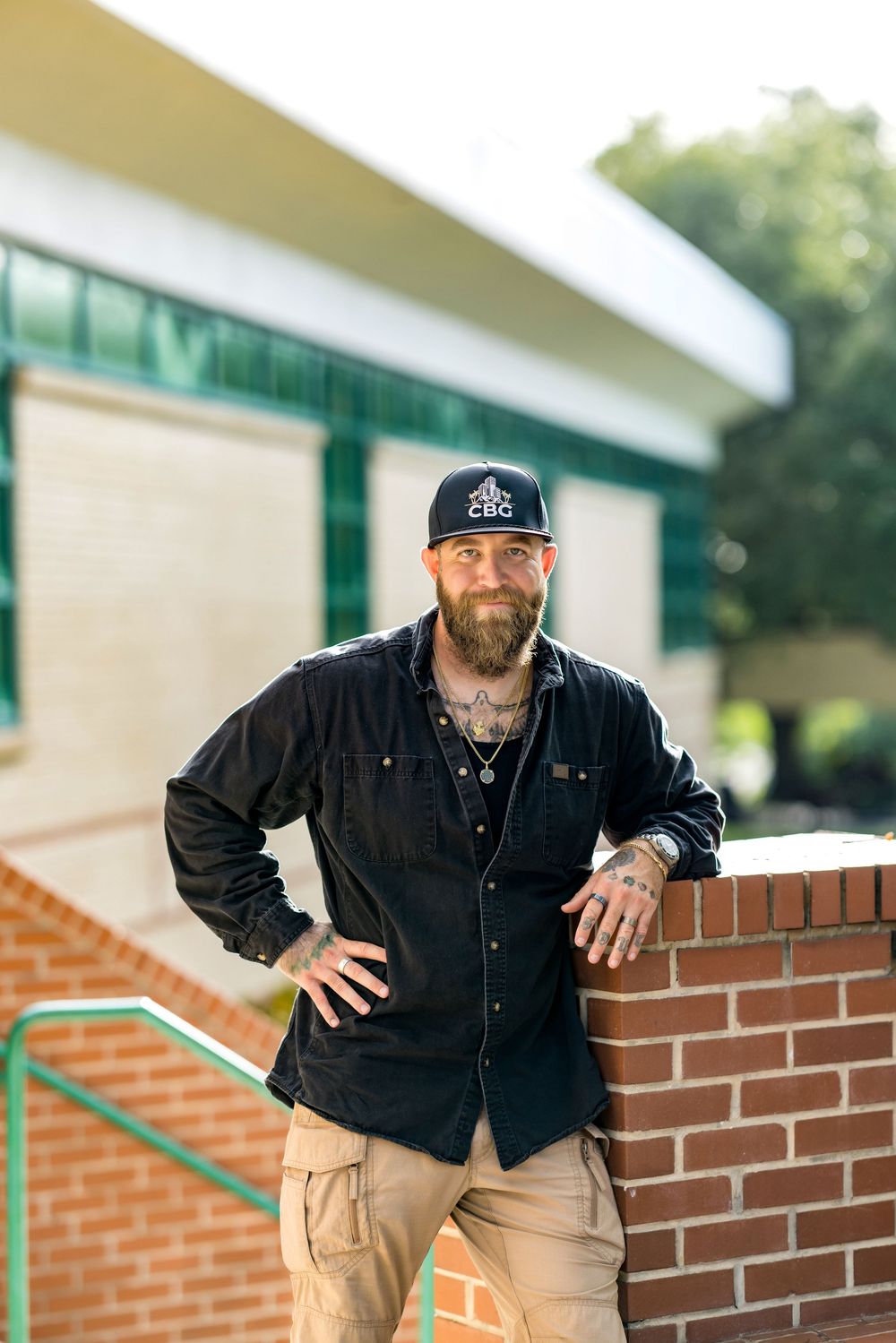 Bearded tattooed man in black shirt and CBC cap leans against brick wall outdoors near green building.