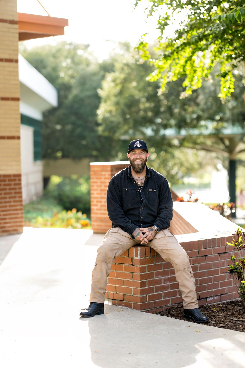 Smiling bearded man in black shirt and cap sitting on brick wall outdoors near building with trees.