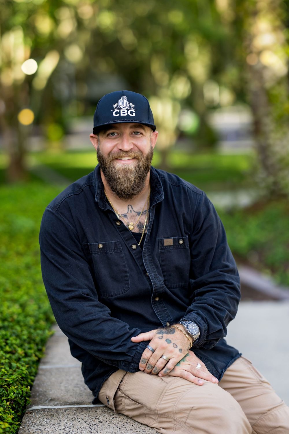 Bearded man with tattoos wearing a navy CBG cap and dark shirt sits smiling outdoors in a lush green park.