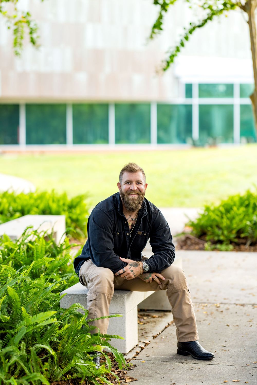 Bearded man in black jacket sits smiling on a concrete bench outdoors, surrounded by lush green ferns near a modern building.