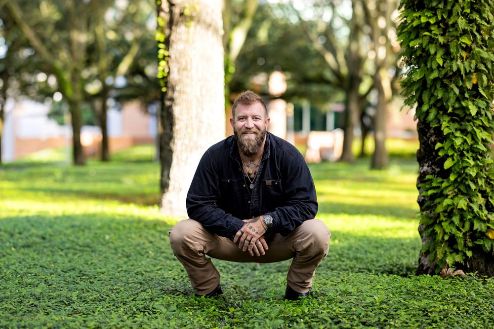 Bearded tattooed man squatting outdoors on green grass between trees, wearing dark jacket and khaki pants.