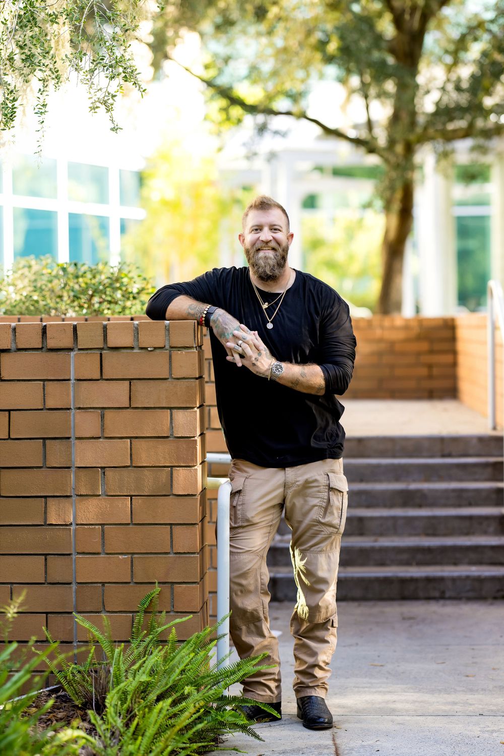 Tattooed bearded man in black shirt and cargo pants leaning on brick wall outdoors, smiling confidently.