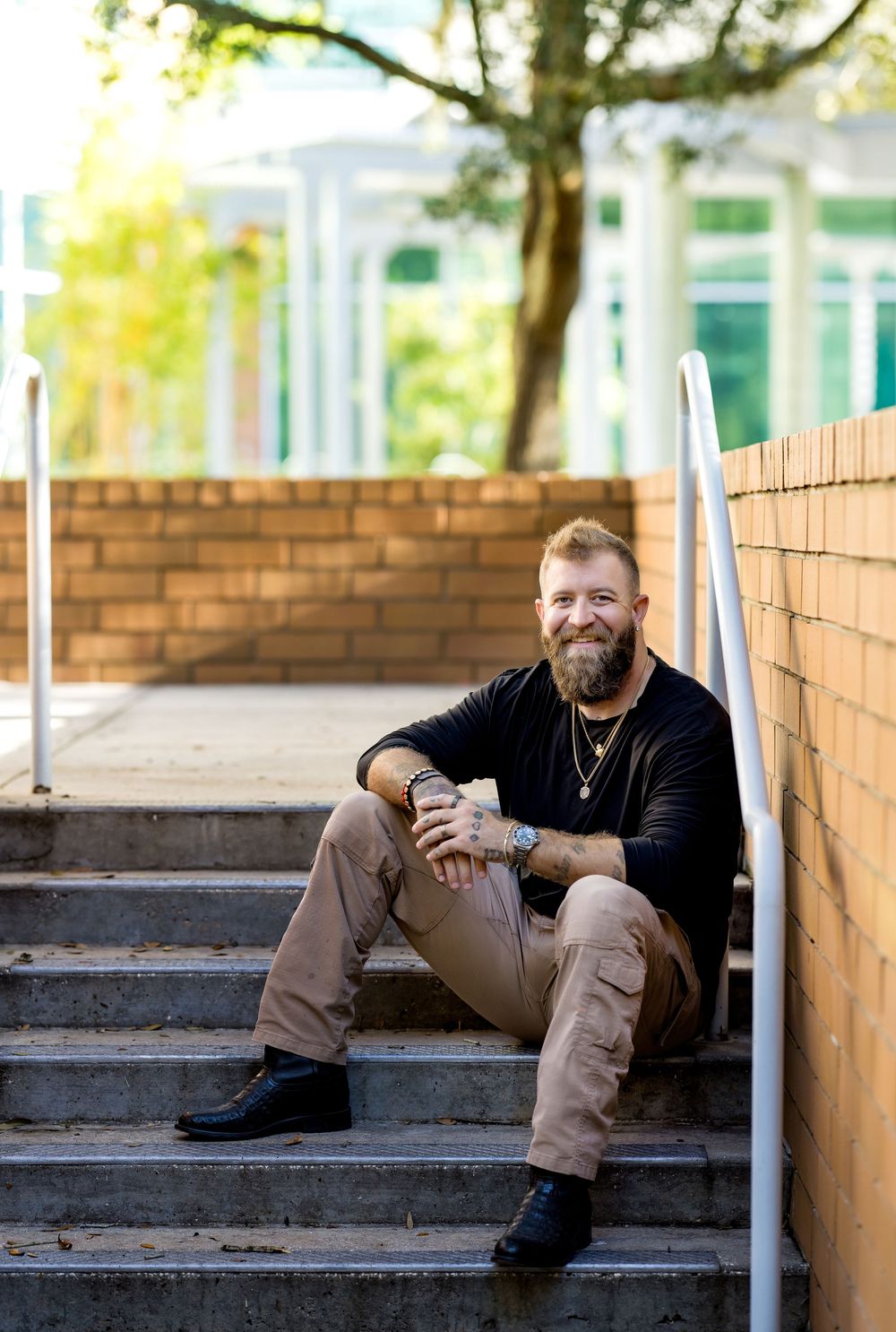 Bearded man with tattoos sitting on outdoor steps, smiling, wearing black shirt and khaki pants.