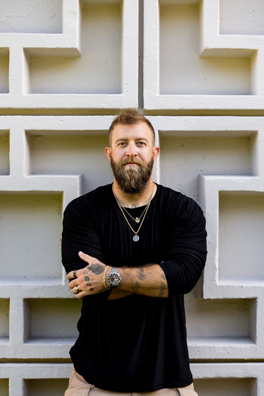 Bearded tattooed man in black long sleeve shirt with necklace and watch, arms crossed, leaning against geometric wall.