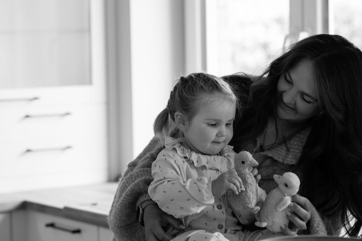 Black and white photo of a mother and young daughter playing with stuffed animal toys near a bright window.