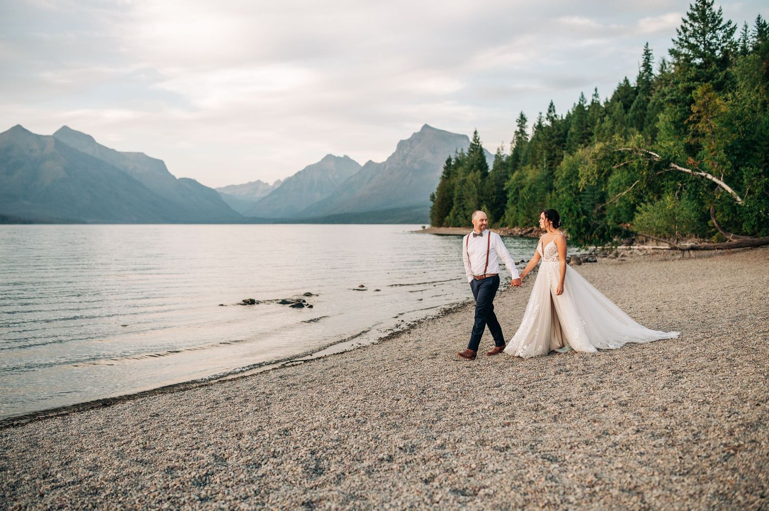 A bride and groom walk hand-in-hand along a rocky lakeshore with mountain and forest backdrop.