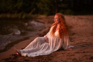 Beautiful redhead woman in a white lace dress sits relaxed on a sandy beach at golden hour.