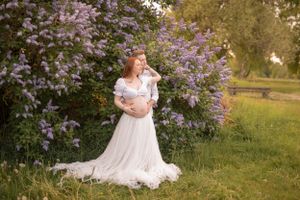 Pregnant woman in an elegant white gown poses gracefully in front of blooming purple lilac bushes.