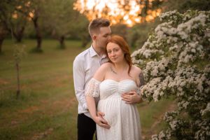 Couple embraces tenderly near white blossoming trees during a romantic golden hour maternity session.