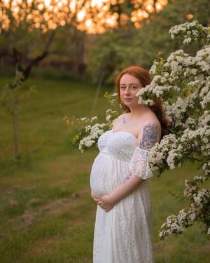 Gravid kvinna står i en vit långklänning och håller om sin mage. Det är grönska och solnedgång i bakgrunden.