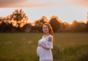 Pregnant redhead woman in a white off-shoulder lace gown cradles her bump in a glowing sunset field.