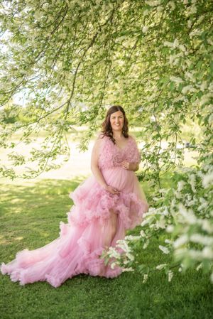 Pregnant woman in a stunning pink ruffled tulle gown poses under flowering white tree branches outdoors.