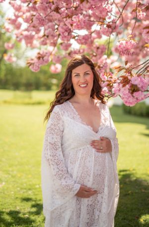 Pregnant woman in white lace dress smiling under pink cherry blossom tree in sunny park.