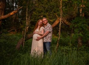 Expecting couple embraces tenderly in a lush green forest during a warm sunset maternity photoshoot.