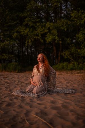 Gravid kvinna sitter på knä på en sandstrand och blickar ut över solnedgången