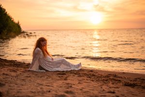Redheaded woman in a white lace dress sits peacefully on a sandy beach during a stunning ocean sunset.