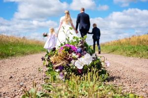 A colorful wildflower bouquet sits on a gravel path as a wedding family walks away under blue skies.