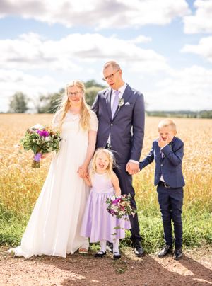 Happy wedding couple poses with two young children in front of a golden wheat field on a sunny day.