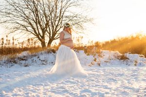 Pregnant woman in white skirt embracing belly near bare tree in snowy winter landscape.