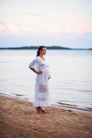 En gravid kvinna har en vit klänning på sej. Hon håller om magen och blickar ut över vattnet där hon står på en strand.