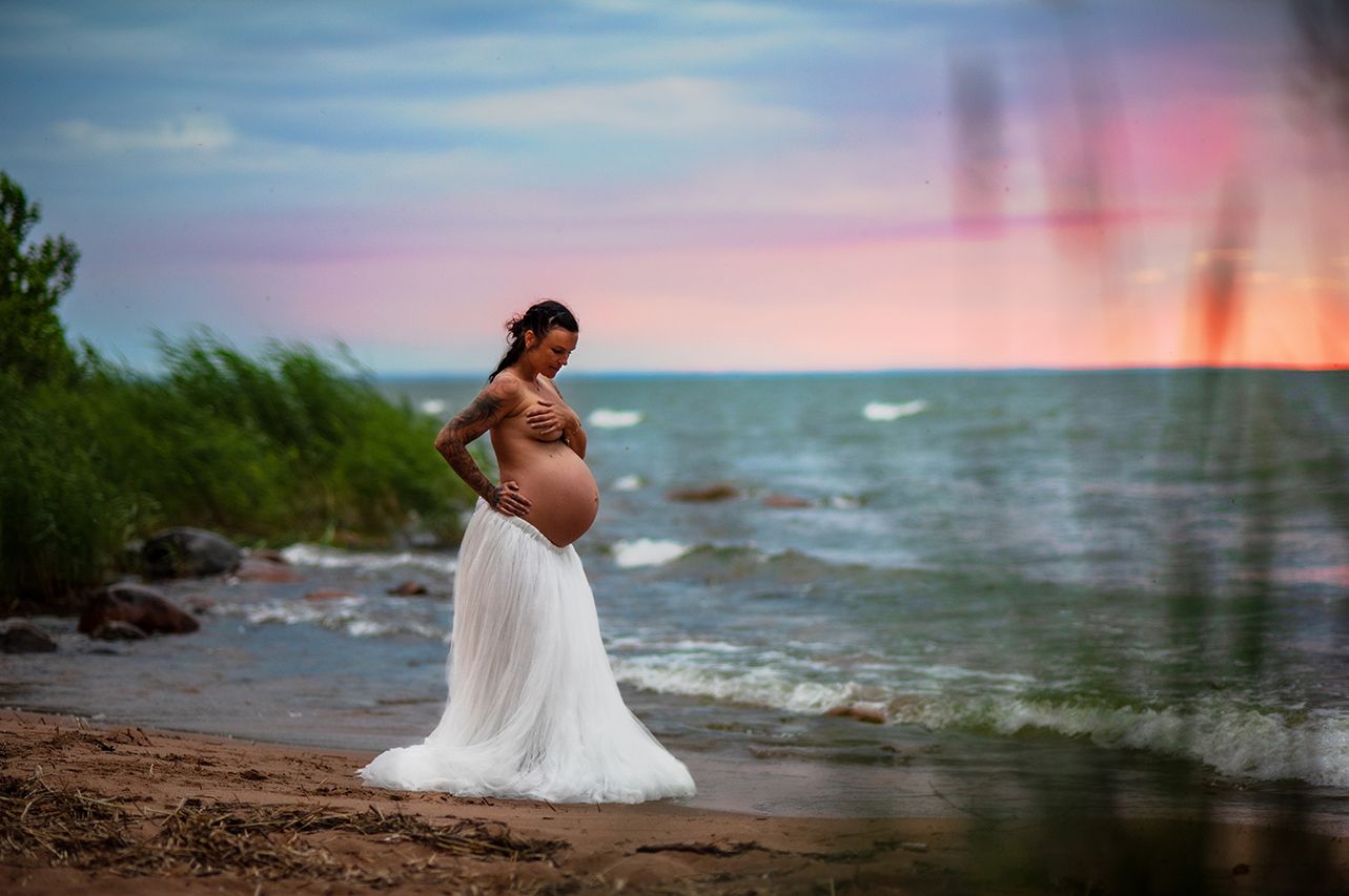 Pregnant woman in white tulle skirt posing on rocky beach shore at colorful dusk.