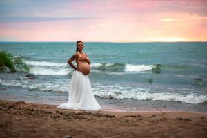 Vacker gravid kvinna står på en strand endast iförd en vit fluffig kjol. Vågorna skvalpar bakom henne och himlen är alldeles rosa.