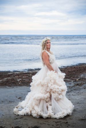Pregnant woman in ruffled white gown with floral crown posing on a rocky beach with ocean backdrop.