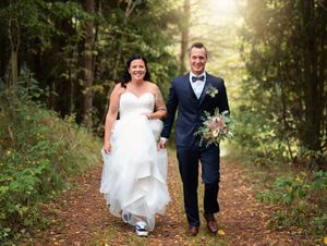 A smiling bride and groom walk hand-in-hand along a forest path, groom holding a bouquet.