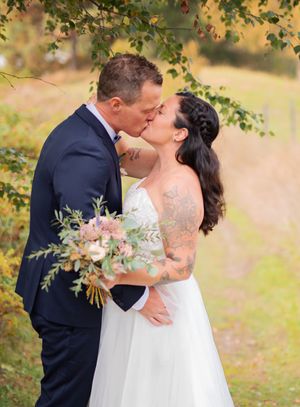 A tattooed bride and groom share a romantic kiss outdoors amid colorful autumn foliage.