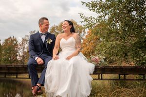 A laughing bride and groom sit together on a bridge surrounded by lush autumn trees.