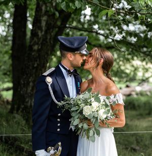 A bride and groom in military uniform share a kiss outdoors beneath a large shady tree.