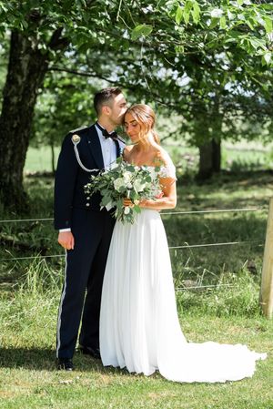 A groom in military dress uniform embraces his bride holding a white bouquet under green trees.