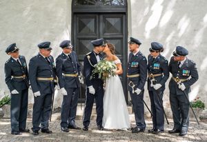 A bride and groom kiss surrounded by six uniformed military officers outside a historic stone building.