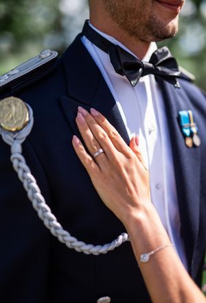 A bride's hand with a wedding ring rests on the chest of a groom in military dress uniform.