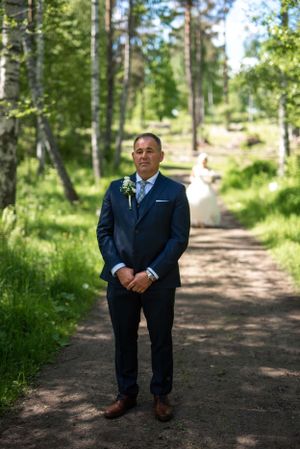 Groom in navy suit with boutonniere stands on forest path as bride approaches in background.