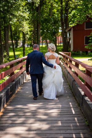 Bride and groom walking hand in hand across wooden bridge toward red building on sunny day.