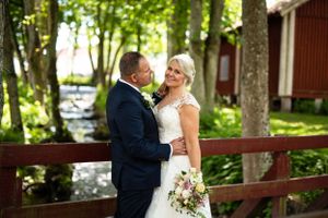 Wedding couple embracing on bridge surrounded by lush green trees near a red building.