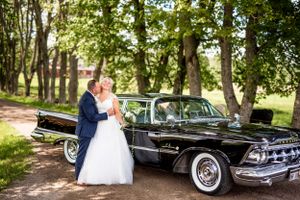 Bride and groom posing beside a classic black vintage car under green trees on wedding day.