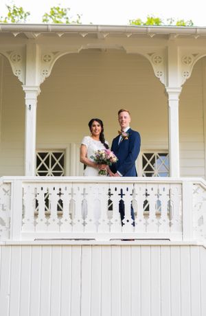Smiling bride and groom standing together on white Victorian balcony holding pink bridal bouquet.