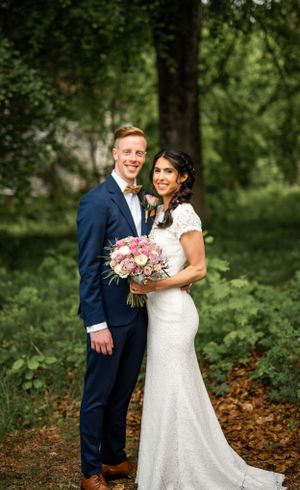 Smiling bride and groom posing together in forest setting with pink and white bridal bouquet.