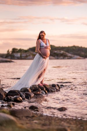 Pregnant woman in a white flowing gown stands on coastal rocks during a golden sunset over calm waters.